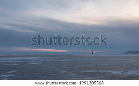 On the endless surface of the frozen lake, cracks and areas of snow are visible. In the distance, against the backdrop of a cloudy sunset sky, tiny silhouettes of people walking on the ice. Baikal