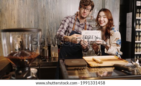 Business owner couple young Asian woman in apron hanging we're open sign on front door  welcoming clients to new cafe. Happy waitess with protective face mask holding open sign at cafe .