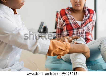 Close up of afro woman doctor examining patient with knee problems in clinic. Mixed race 10-aged girl at the appointment of doctor orthopedist at modern medical center Royalty-Free Stock Photo #1991350151