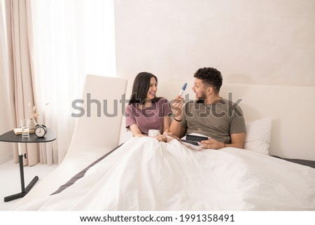 Happy woman looking at her surpwised husband with pregnancy test while lying in bed