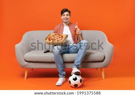 Young excited man football fan in shirt support team with soccer ball sit on sofa at home watch tv live stream eat pizza drink beer isolated on orange background People sport leisure lifestyle concept