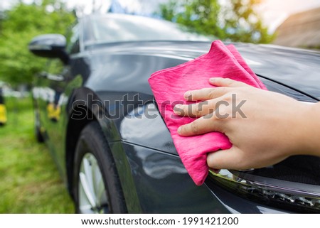 A hand with a red rag wipes water drops on a car after washing with car shampoo and wax in the country. Hand wash