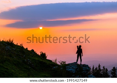 The girl stands on top in a delicate black dress at dawn with arms raised. In horizontal orientation