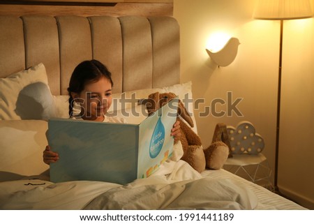 Little girl reading book in bedroom lit by night lamp