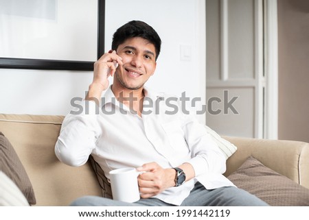 Latin man in white shirt on sofa holding mug while talking on smartphone