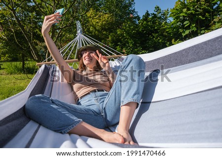 Spanish young woman with bucket hat lying in hammock outdoors taking a photo of herself.