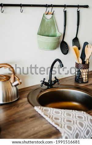 A kettle on the countertop with household utensils. Aluminum sink with a towel on the edge.