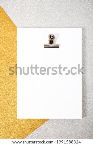 White foil packet and paper clip on a silver and gold textured background. The photo has a copy space, it is taken from an overhead point of view and is in vertical format.