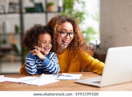 Young positive mother with happy son sitting at desk and usinng laptop, looking at screen and smiling while watching funny videos together. Happy afro american family spending leisure time at home