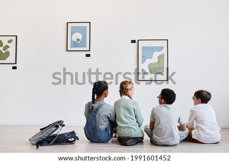 Back view at diverse group of children sitting on floor in modern art gallery and discussing paintings, copy space