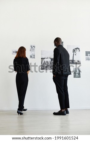 Minimal back view at two people wearing black while looking at photographs in modern art gallery