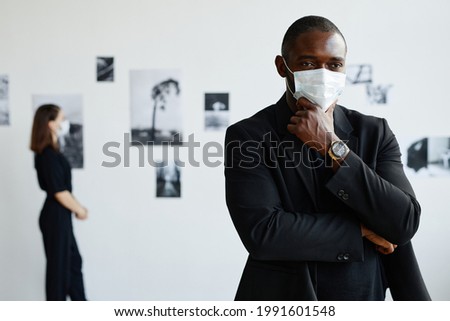 Minimal waist up portrait of elegant African-American man wearing mask while looking at photographs in modern art gallery, copy space