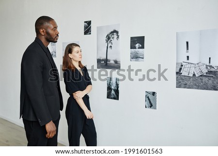 Portrait of elegant mixed-race couple wearing black while looking at photographs in modern art gallery, copy space