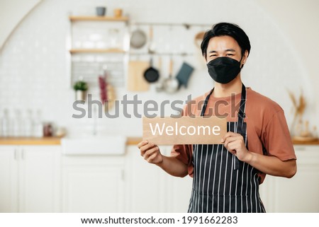 portrait of young asian men waitress wearing a surgical mask hanging a sign that says welcome with restaurants background. Small business owner with face mask reopening the place after the quarantine.