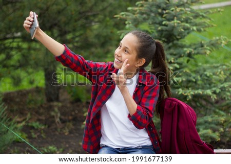 A cheerful girl with long dark hair in a white T-shirt and a plaid shirt in the park uses the phone to communicate in the messenger take a selfie and show the victory sign. Selective focus.