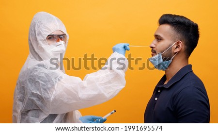 Health worker in protective suit taking Nasal swab from a potentially infected young guy. Corona PCR Virus Test With Swab concept. Yellow background studio. High quality photo. 