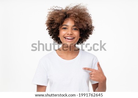 Laughing african american girl showing her blank white t-shirt with finger, copy space for your logo, isolated on gray background