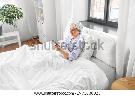 technology, old age and people concept - senior woman with smartphone and wireless earphones in bed at home bedroom