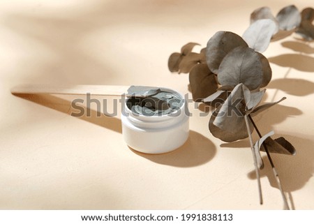 beauty, cosmetics and object concept - blue cosmetic clay mask in jar with wooden spatula and branch of eucalyptus populus on beige background