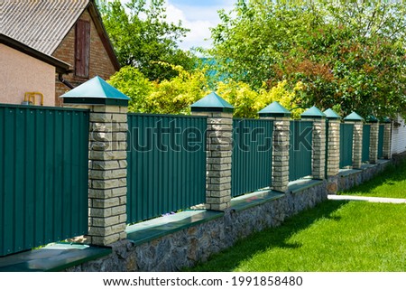 Green metal corrugated fence with brick pillars on the background of a beautiful house. Royalty-Free Stock Photo #1991858480