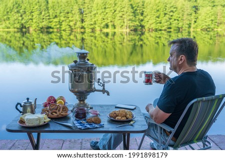 Man drinking tea by the lake. Vintage metal tea samovar with white smoke and food on the table near the calm water lake in green forest at morning, Ukraine