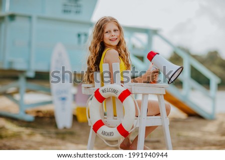 Young pretty curly girl sitting on high white chair with lifeline and posing with megaphone on the beach against blue lifeguard tower.
