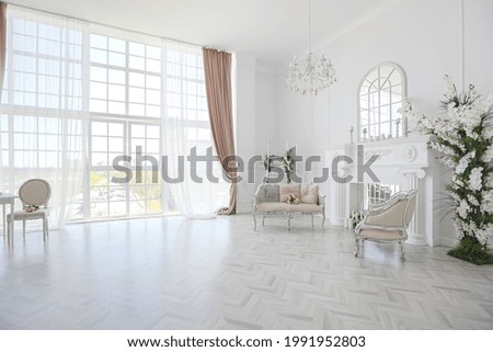 luxurious bright spacious guest room with beautiful chic furniture a huge floor-to-ceiling window in a royal style is decorated with green plants, white walls with stucco and a fireplace
