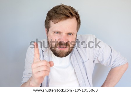 Irritated, disgruntled bearded man looks evil and threatening gesture with his finger. Gray background.