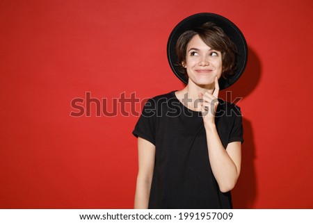 Pretty pensive funny young brunette woman 20s years old wears casual basic black t-shirt hat stand put hand prop up hand on chin look aside area isolated on bright red color background studio portrait
