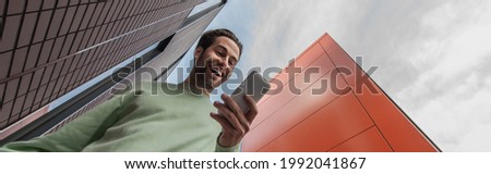 low angle view of amazed man in sweatshirt holding smartphone near buildings, banner Royalty-Free Stock Photo #1992041867