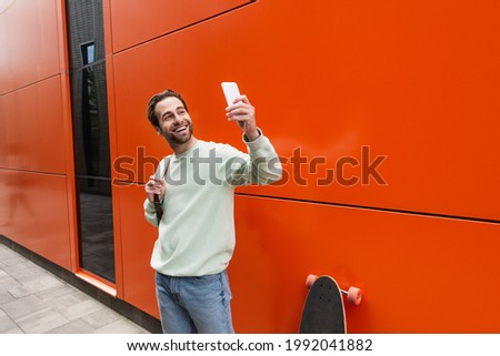 cheerful man in sweatshirt taking selfie on smartphone near orange wall and longboard