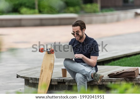 happy man in sunglasses and polo shirt using laptop near smartphone, paper cup and longboard