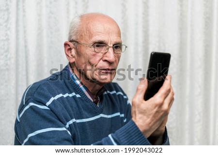 Serious senior man wearing glasses standing at home against white background and using smartphone and looking at camera