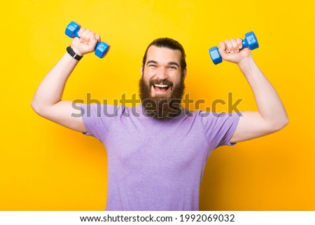Portrait of happy cheerful bearded man working with small dumbbells over yellow background. Royalty-Free Stock Photo #1992069032