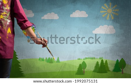 Close up artist hand holding paintbrush. Painter in shirt standing on background colorful picture. Summer landscape with green field and blue sky artwork. Creative hobby and profession.