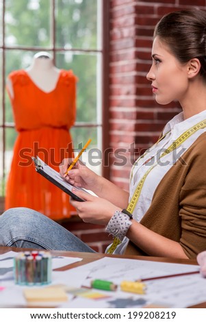 Young and full of creativity. Side view of beautiful young woman drawing fashion sketch and looking away while sitting at her working place with mannequin standing in the background