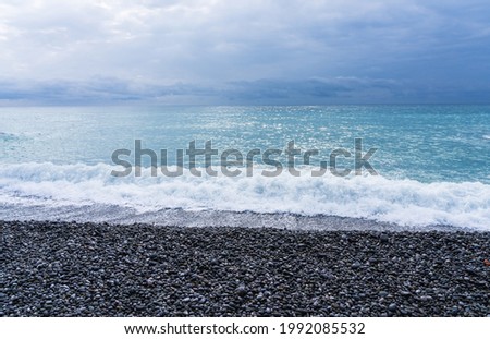 A white wave washes the round pebbles on the beach and the turquoise sea reflects the light of the sun. Photo in calm pastel colors