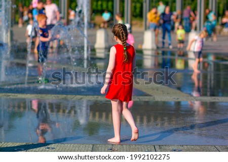 A girl in a red dress walks barefoot by the fountain on a hot sunny summer day in the city. Sultry heat on city streets, family vacation. Royalty-Free Stock Photo #1992102275