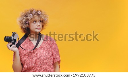 Attractive young photographer isolated on bright yellow background studio. Camera hanging from her neck. Dressed in casual pink top and wearing flowers in her hair. Leisure Photographer Concept. 