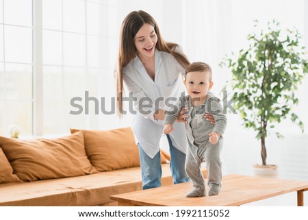 Young single mother teaching her little son toddler baby infant how to walk, crawling on the table, making first steps at home. Motherhood and childcare, walking exercises for kids and children. Royalty-Free Stock Photo #1992115052
