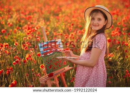 Portrait of Little girl in the field of red poppies On the Sunset and painting on the canvas placed on a drawing stand, smiling and looking at camera. Be a part of learning outside of the school.