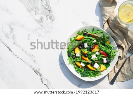 Fresh homemade salad with nectarines, blueberries, arugula, spinach and feta cheese on white marble background with hard shadows. Healthy vegetarian food. Top view. Space for text. Royalty-Free Stock Photo #1992123617