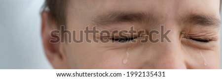 Teardrops rolls down the boy's cheeks, his eyes are closed, he is upset and crying. Sad, unhappy emotions of child, banner size. Royalty-Free Stock Photo #1992135431
