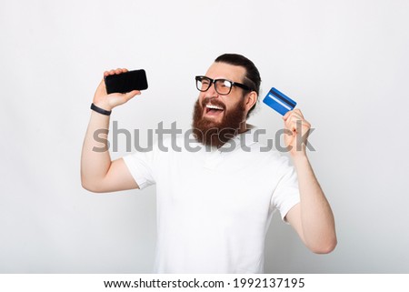 Portrait of excited bearded man holding smartphone and blue credit card.