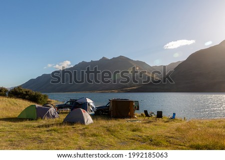A photo of a group of tents put up by the lake. Two unrecognizable cars standing sideways behind the tents. Mavora Lake and Livingstone Mountain range in the background. Sunny, summer day before sunse Royalty-Free Stock Photo #1992185063