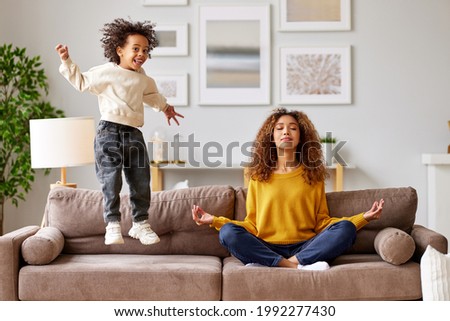 Young calm african american mother in casual wear sitting in lotus pose on floor in living room and meditating while little boy active son jumping on sofa and playing around. Mindful parenting concept Royalty-Free Stock Photo #1992277430