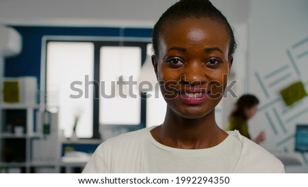 Close up of african woman looking at camera smiling standing in start up creative agency office holding laptop, typing on it. Black videographer editing video project using post production software