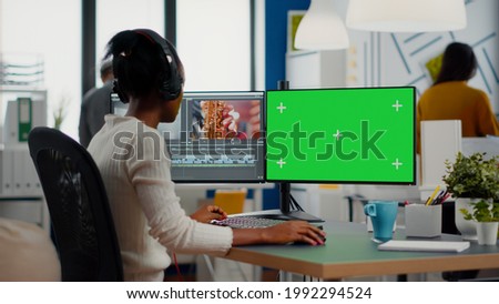African woman videographer with headset processing film montage working at greenscreen, chroma key isolated display of computer. Video editor editing movie in post production software in agency office