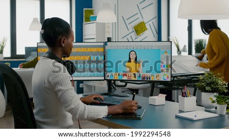African woman retoucher sitting in modern creative photo studio working on computer after photoshoot, retouching photographs of model in image editing software with stylus pencil, color greading app.