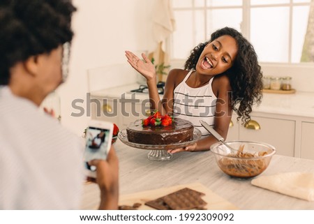 Happy young girl standing in kitchen with cake being photographed by her mother. Daughter with chocolate cake at home with her mother taking her picture using mobile phone.